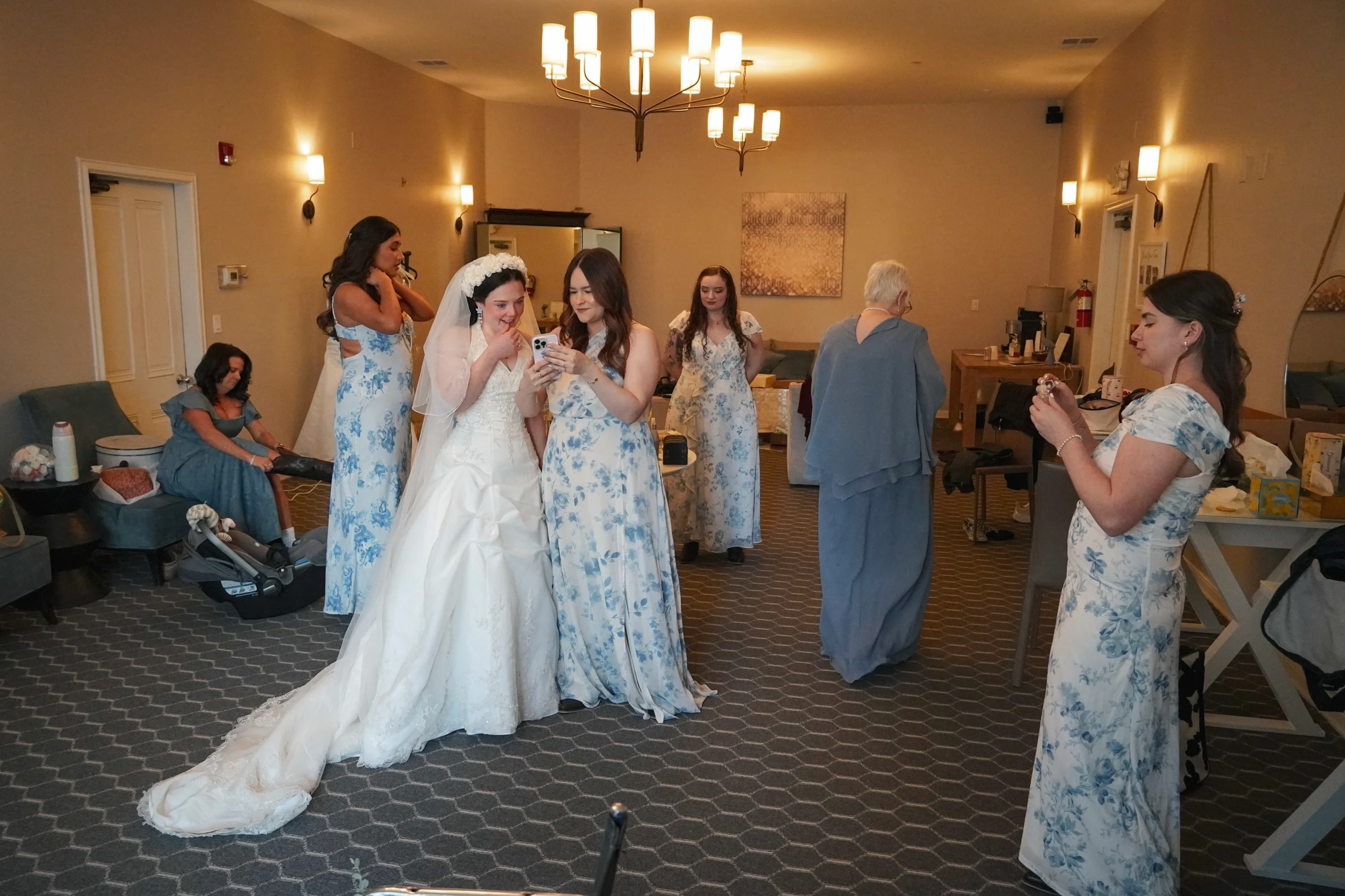 A candid photograph of the girls getting ready for the wedding ceremony. One of the bridesmaids is showing the bride something on her phone, and the bride is smiling. The brides dress is flowing perfectly down her side. The other women in the room are putting on their last minute touches like shoes and makeup. The lighting in this photo is warm and dreamy.