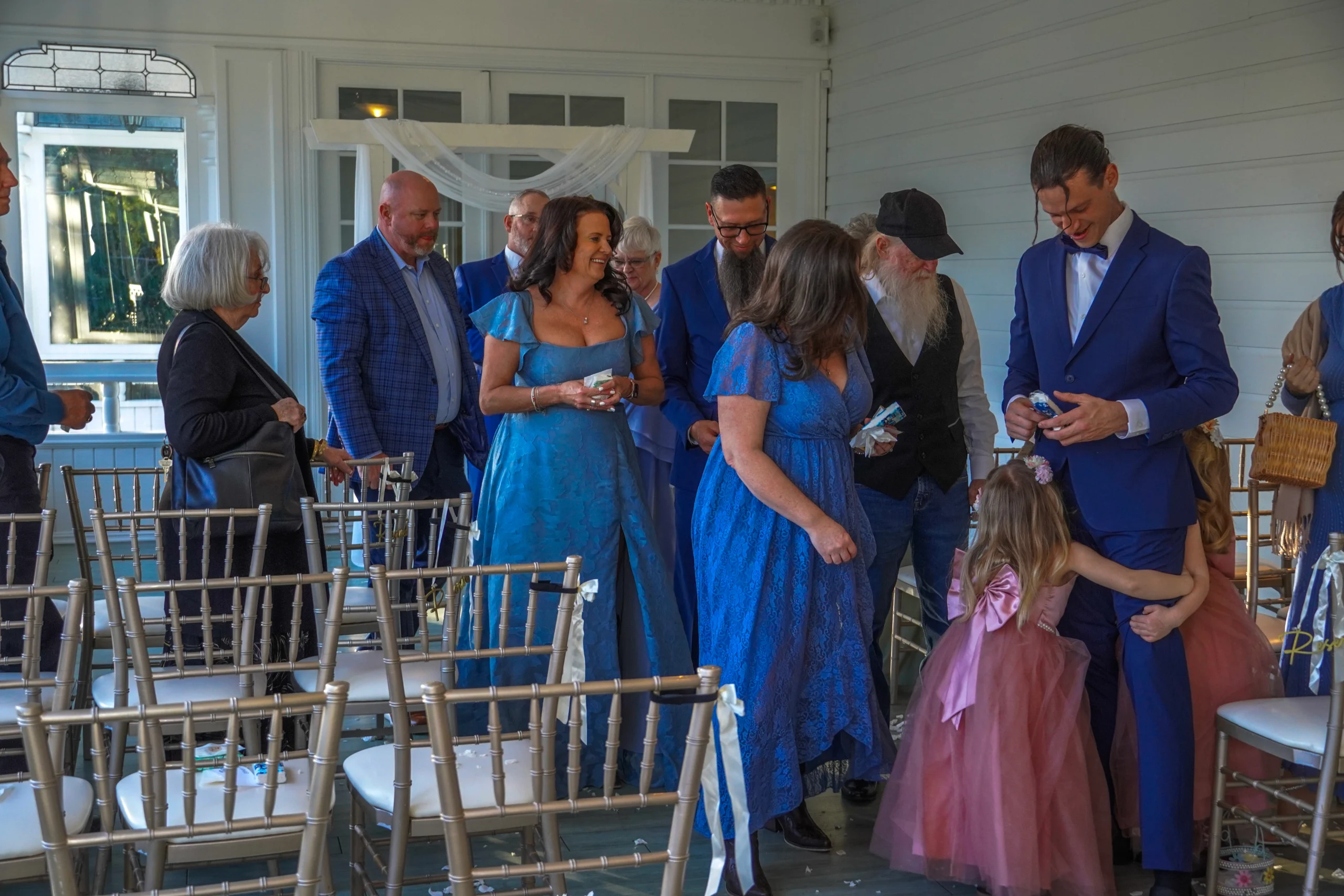 The wedding guests are exiting the ceremony while smiling. It looks like they enjoyed it. Most people are dressed in blue. The flower girls are in pink dresses with cute bows on the back and hug their dad's legs while he smiles at them. They also have pink flowers in their hair.