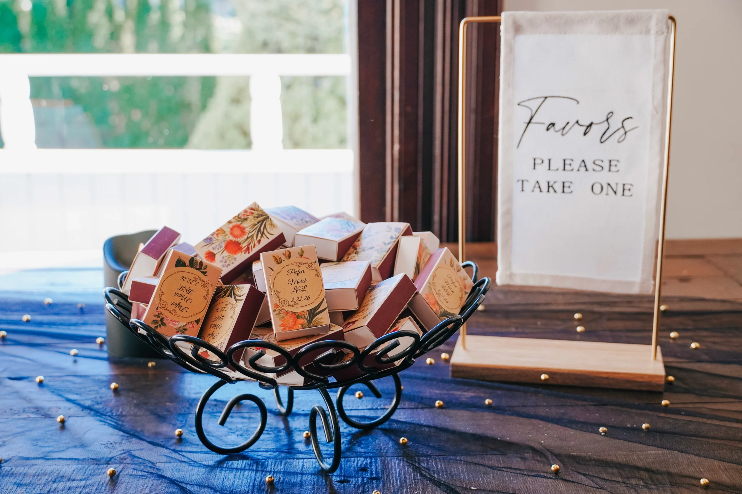 Detail wedding photography of give aways for the wedding guests. There is a sign that says "Favors please take one", a basket of boxes of matches in front of the sign that say "Perfect match" with a window behind the two items, giving the detail photo a light and airy feel.