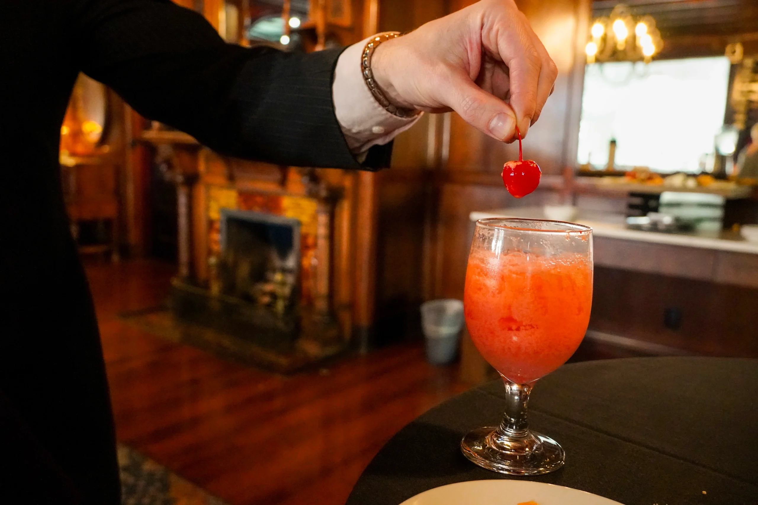 A close up detail image of a man in a black suit with a mocktail that is brightly colored pink / orange. The man is taking the cherry off of the top of the drink, and letting the drink drip off of the cherry. There is a fireplace and warm lighting in the background. It looks very elegant, classy, upscale, and fancy. It is also photographed to be dark and moody.