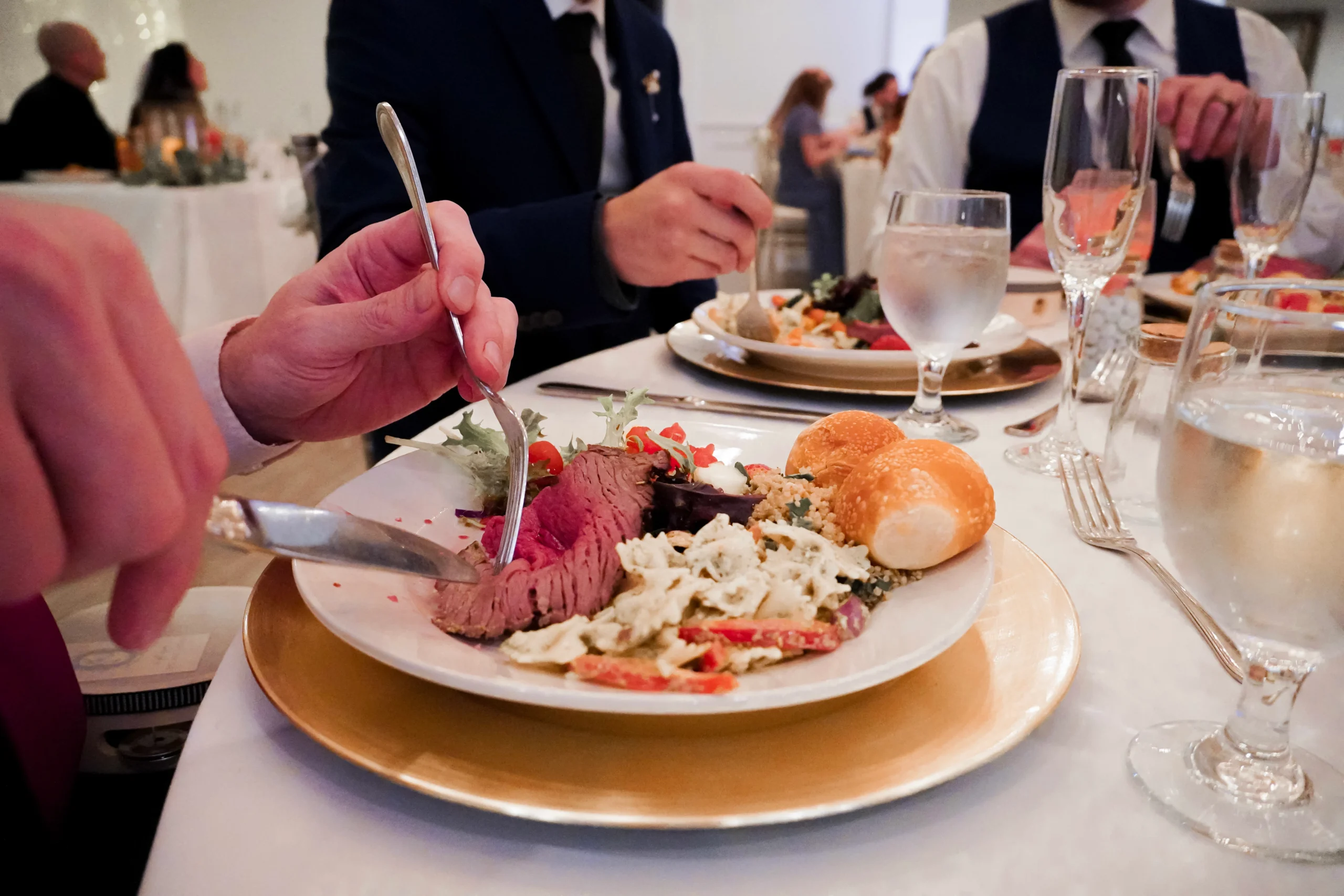Wedding dinner detail photography of best man, groomsmen, and friend eating their dinner. All of them are holding their forks and knives in their hands. A close up image of the plate shows a mix of red meat, pasta, dinner rolls, salad, fruit, and vegetables. There are also water glasses and champagne flutes on the table. The two men in the wedding party are both wearing black ties.