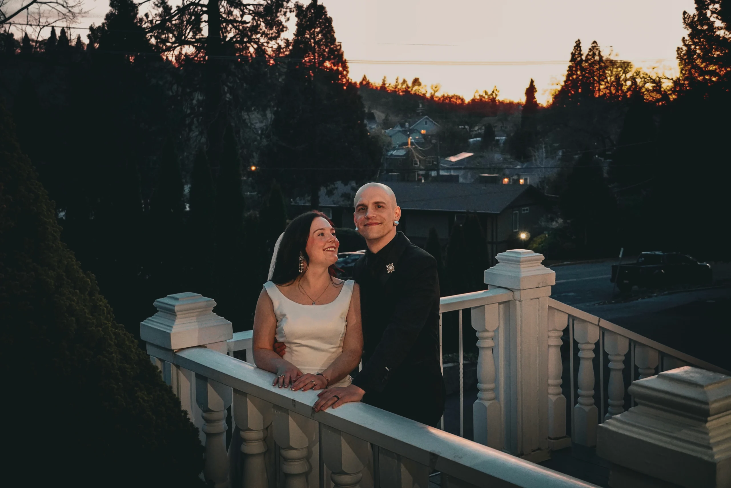 The bride and groom are standing outside on a staircase balcony with white railing under a beautiful orange sunset in the Sacramento, California area. The bride is in her white dress and looking up, smiling at the groom. The groom is in his black suit, smiling at the camera.