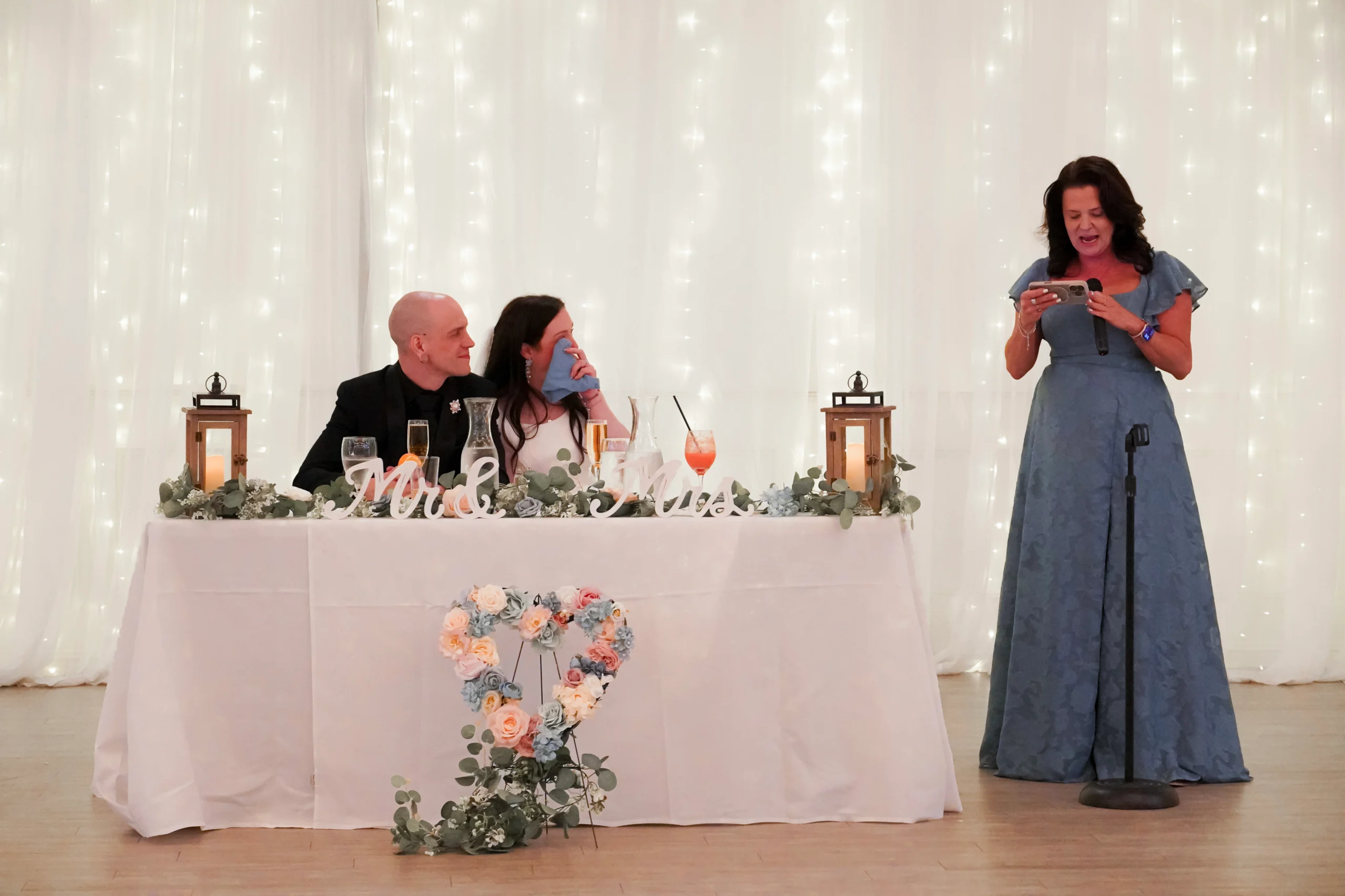 The brides mom makes a speech. The bride is crying and wiping her tears with a napkin. The groom is smiling and sitting close to the bride. The mom is holding a microphone and in the middle of speaking. There are glistening lights behind them.
