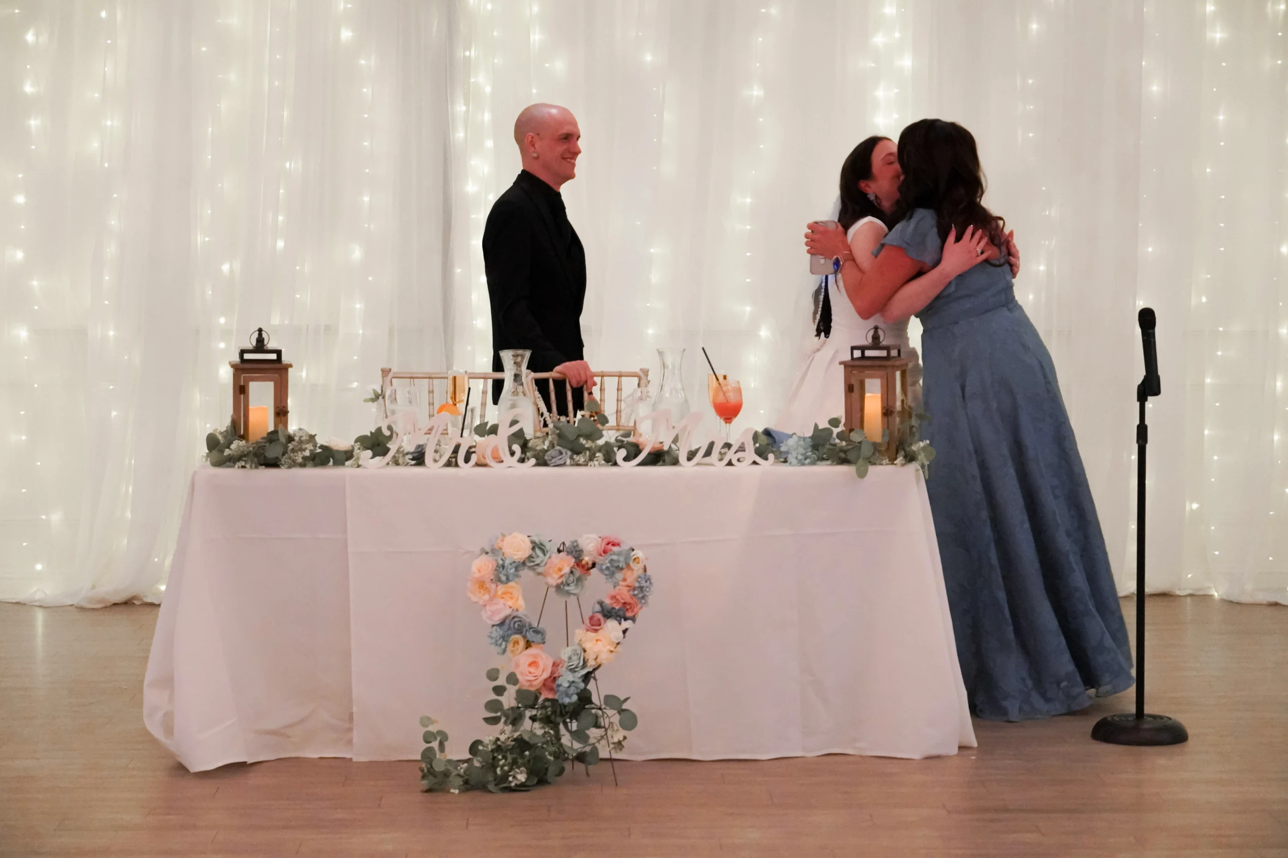 Mother of the bride hugs the bride after giving a speech at the reception dinner. The groom is walking over to them also smiling. There are twinkly lights behind them.