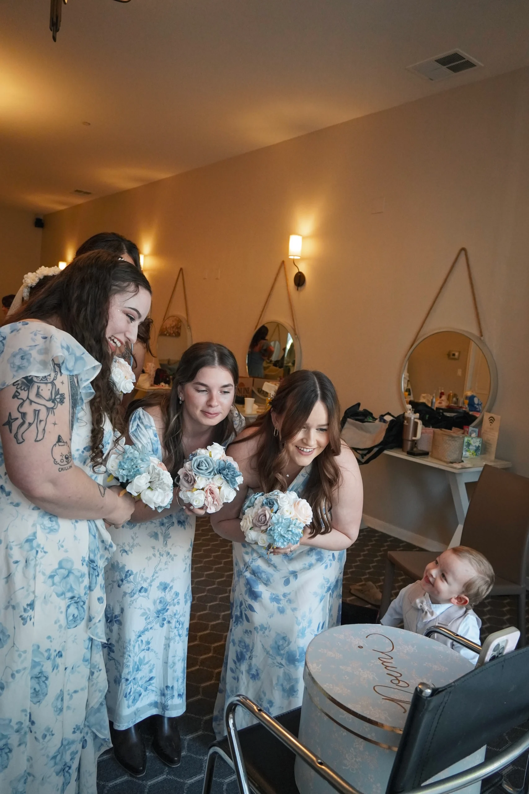 A baby smiles at the bridesmaids while they make content for social media. They are all in floral blue dresses holding bouquets of flowers and smiling in great lighting.