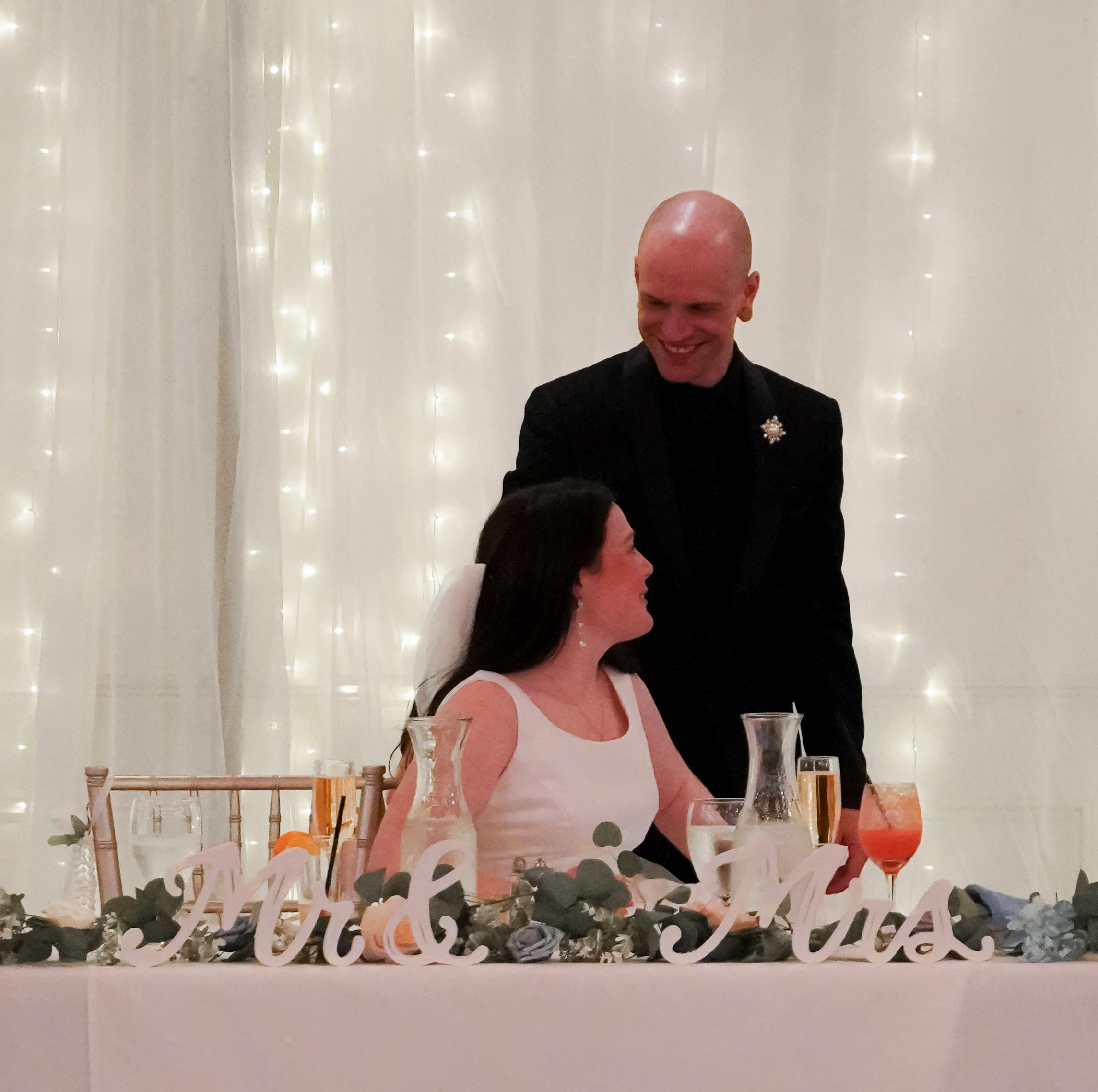 The groom stands behind the bride, who is sitting at their dinner table. She looks up at him, and he smiles at her. The table has their drinks, food, flowers, and says "Mr. & Mrs." in white cursive writing. There are sparkly lights behind them giving the photo a dreamy aesthetic.