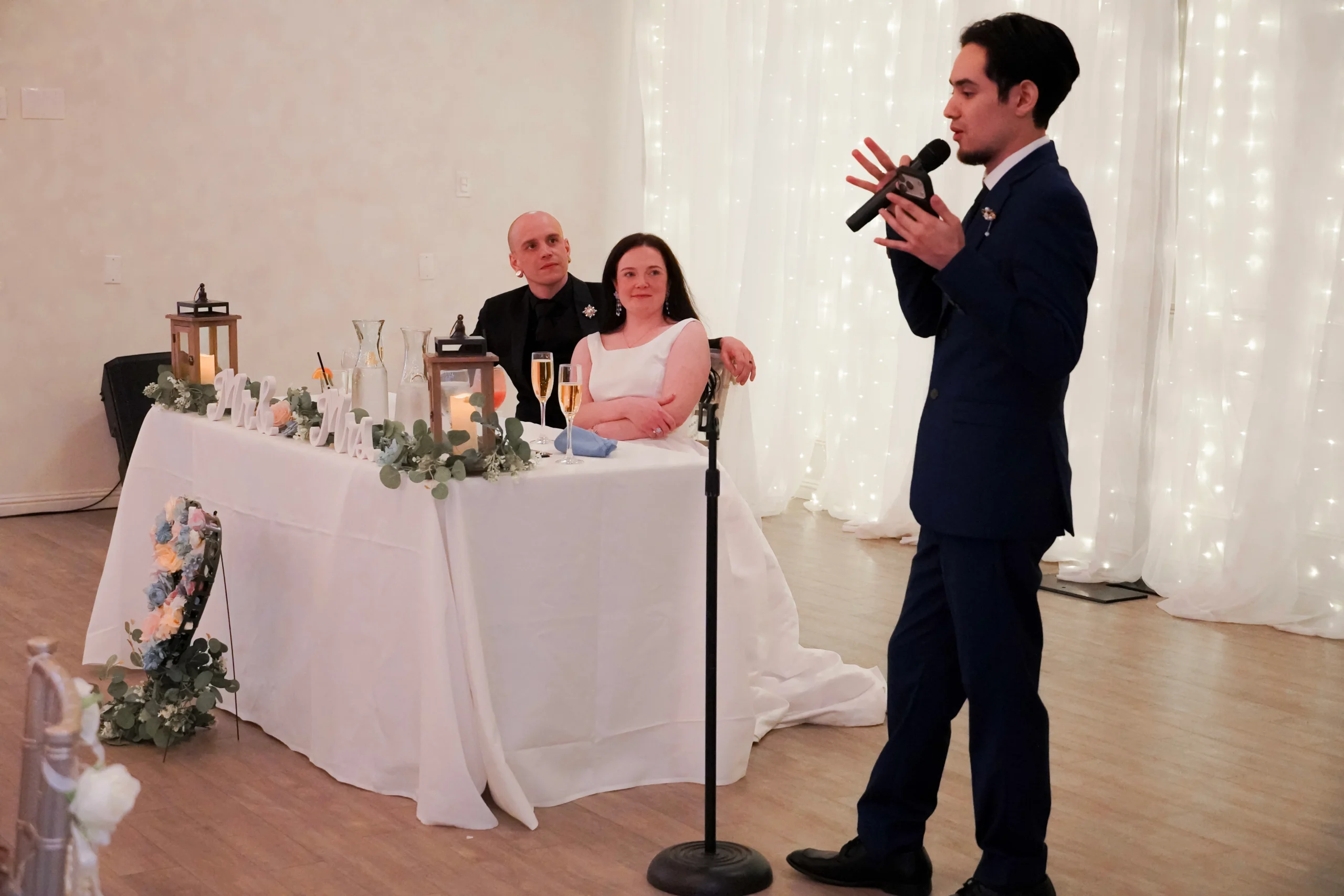 The best man makes a speech at the wedding reception. He is holding a microphone and standing in a pose like he's talking. The groom and bride are sitting at a table next to him while looking at him making the speech. The bride is smiling and the groom looks like he is intrigued. The couple is sitting at their table that says "Mr. & Mrs." with flowers and champagne glasses on it. There are sparkling lights behind all of them.