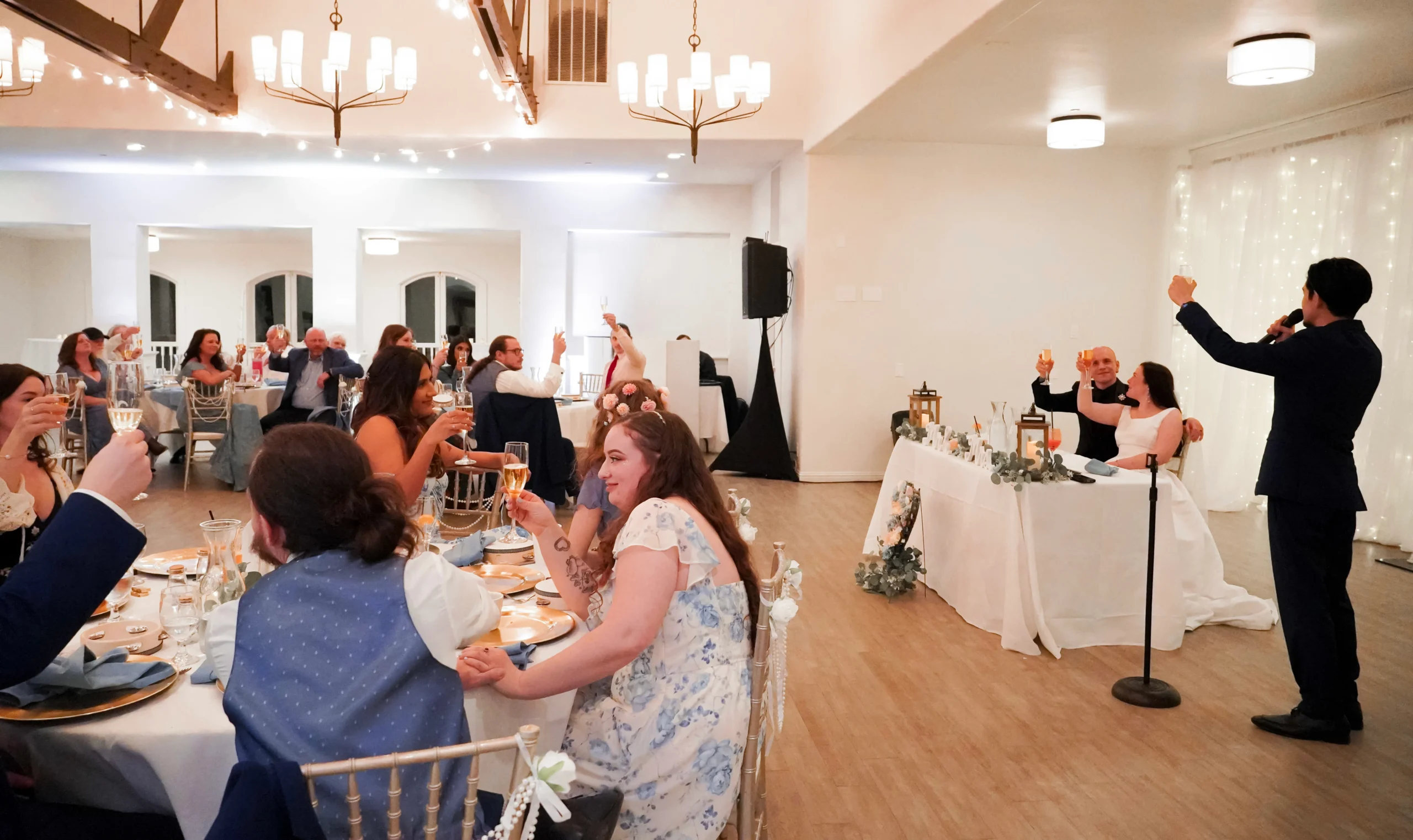 The best man of the groomsmen gives a speech and makes a toast at the wedding dinner. The bride and groom are holding up their glass at a table in front of their friends and family, who are also holding up their flute glasses, or flutes. The room is warmly lit. Most of the guests are dressed in light blue.