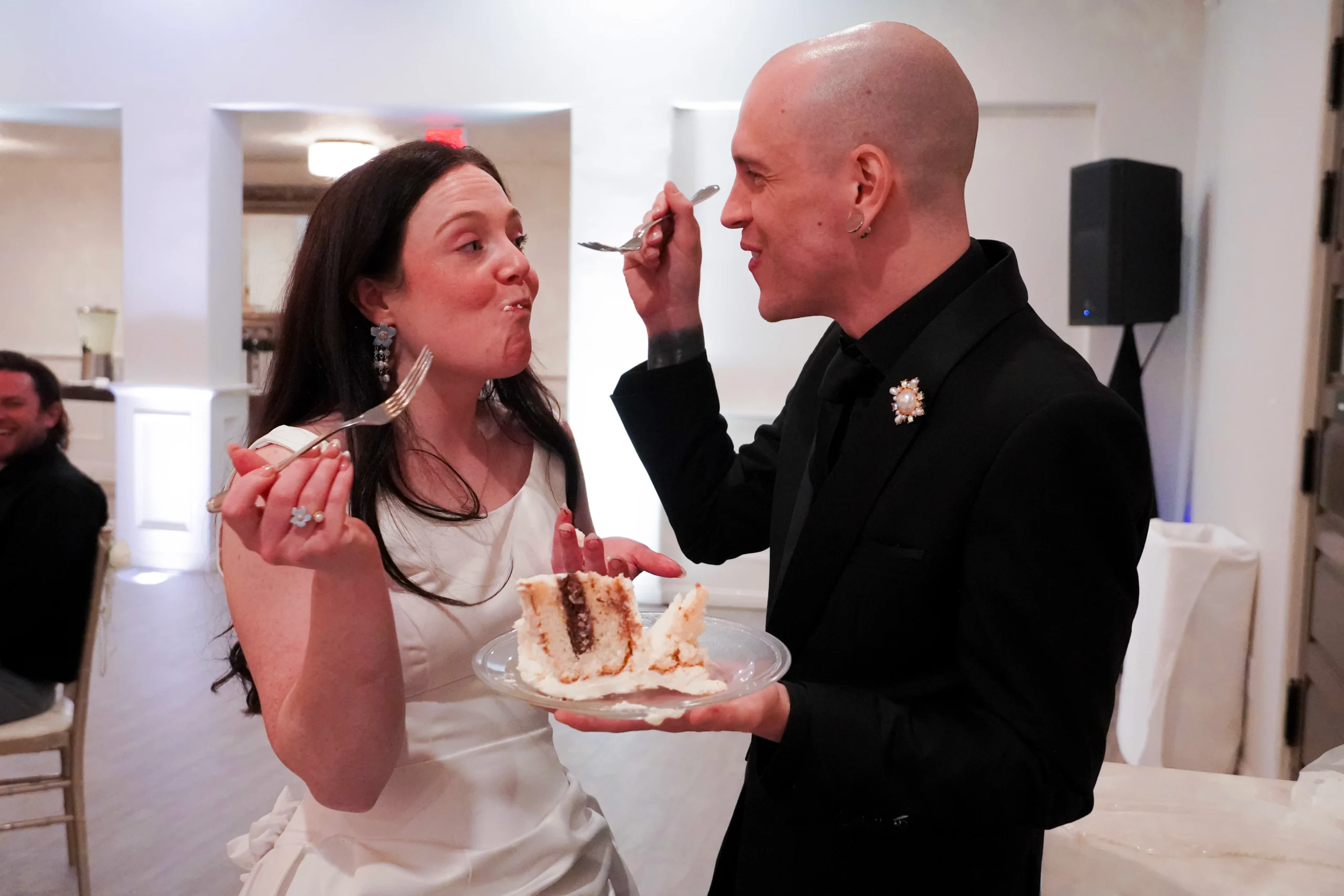 The newlywed couple cut the cake and feed it to each other. The bride is holding up a fork, looking at her groom, with frosting on her lips. The groom is looking at his bride while smiling and holding a slice of cake and a fork. It looks as if they have just fed each other a bite of their wedding cake.