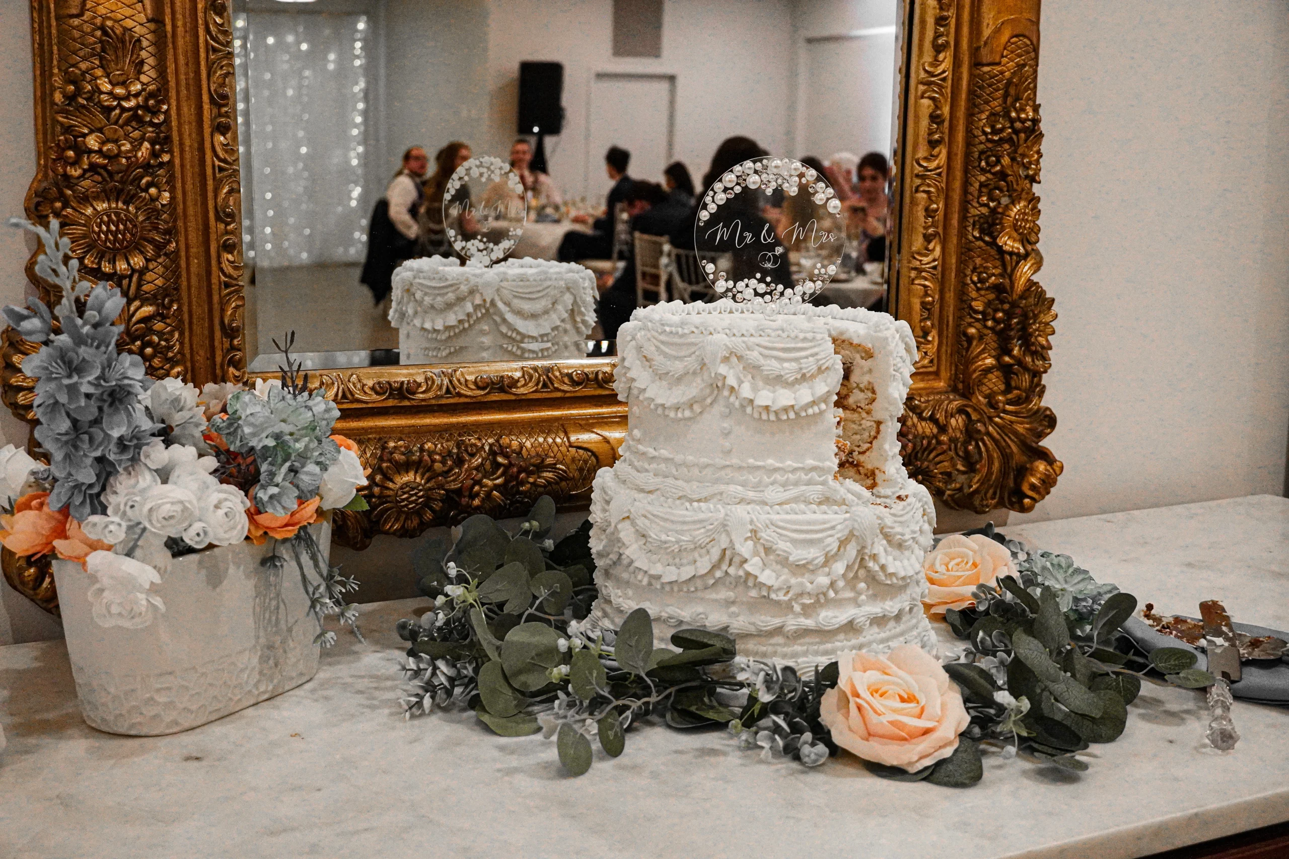 Wedding detail photography of the cut wedding cake Fine art wedding detail photography of the cake after it was cut by the newlywed couple. The elegant tiered cake is white, decorated with white frosting, and has peach colored flowers and green leaves on the table around it. There is one piece cut from the cake. There is a little chocolate center inside the cake. There is a clear sign with pearls on it that says "Mr. & Mrs." on the top of the cake, as a cake topper. There is a mirror behind the cake with a golden frame. The mirror reflection shows the cake and also the wedding guests that are mingling. Also on the table are the cutting utensils used to cut the cake, crossed in a cute way, with cake and frosting on them.