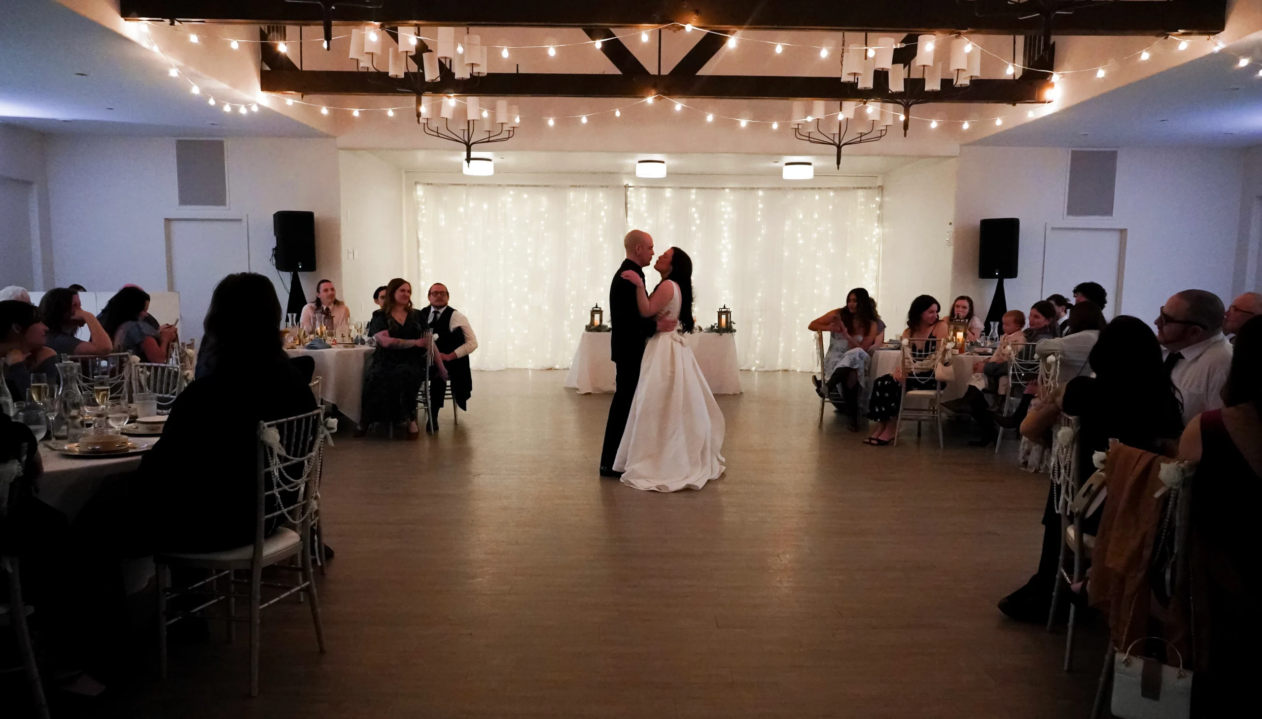 Bride and groom dance their first dance. They are looking at each other, his hands on her waist and her hands on his shoulders, and look like they are about to kiss. They are in the center of the room, with their wedding guests watching them in awe from their dinner tables. The room is dimly lit with twinkling warm white lights above and behind them. It looks like the perfect ballroom wedding photo.
