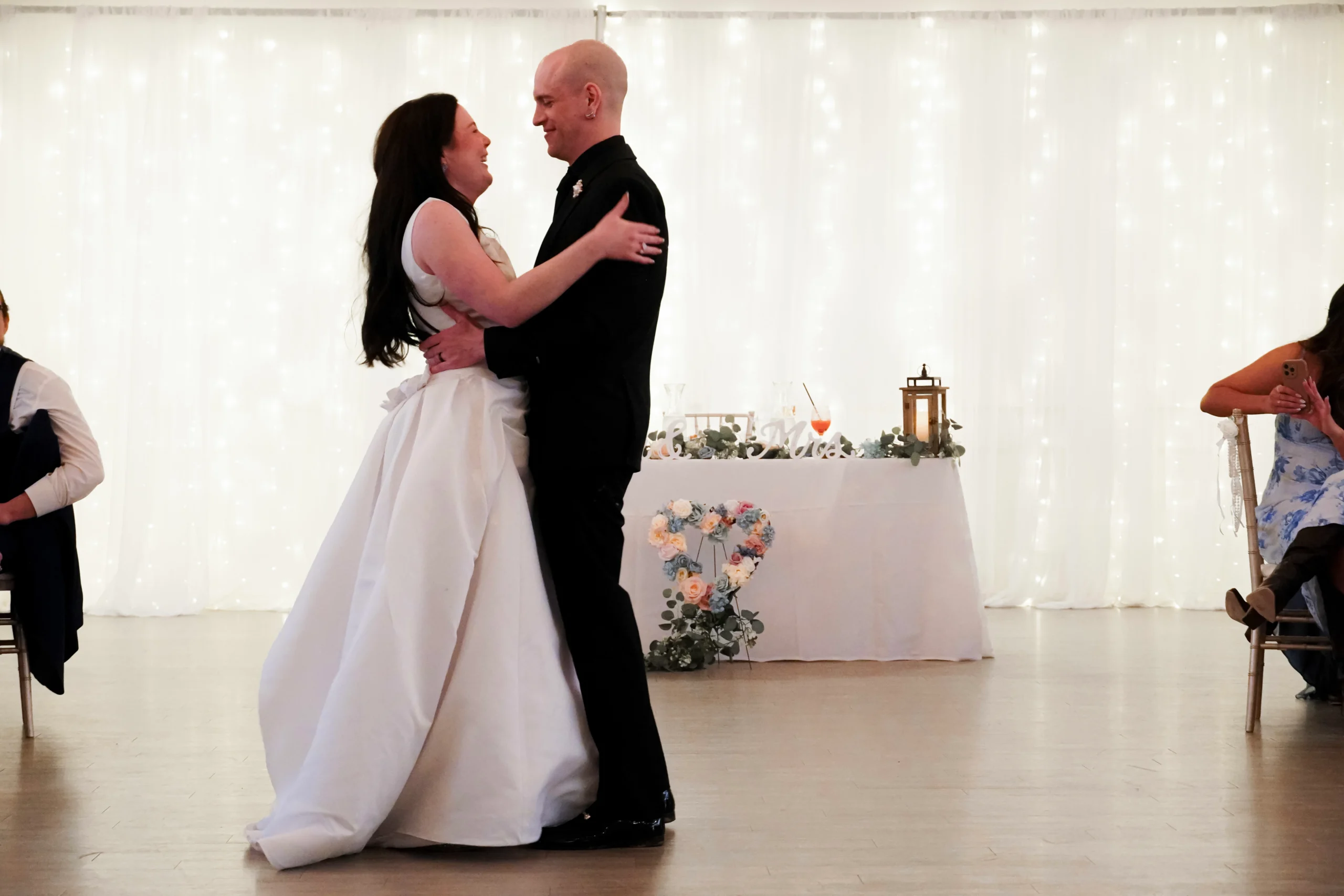 The bride and groom dance their first dance at the wedding reception. They are looking at each other and smiling and laughing. His hands are her waist, and her hands are on his shoulders. Behind them, the warm lighting is shining, there are heart shaped flowers, and their table that says "Mr. & Mrs." has their food and drinks on it.