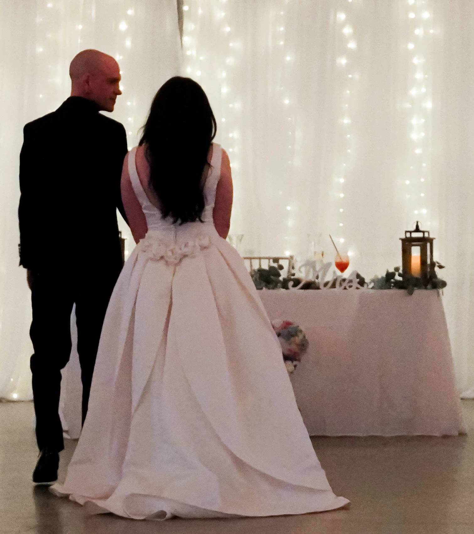 The bride and groom walk back to their table that says "Mr. & Mrs." during the wedding reception. They stop in front of their table while he looks at her and holds her hand, and she shows the back of her beautiful dress. The dress looks like a princess dress. It is white, has an hourglass silhouette, flows out at the bottom, and has white flowers in a v shape below her hair.