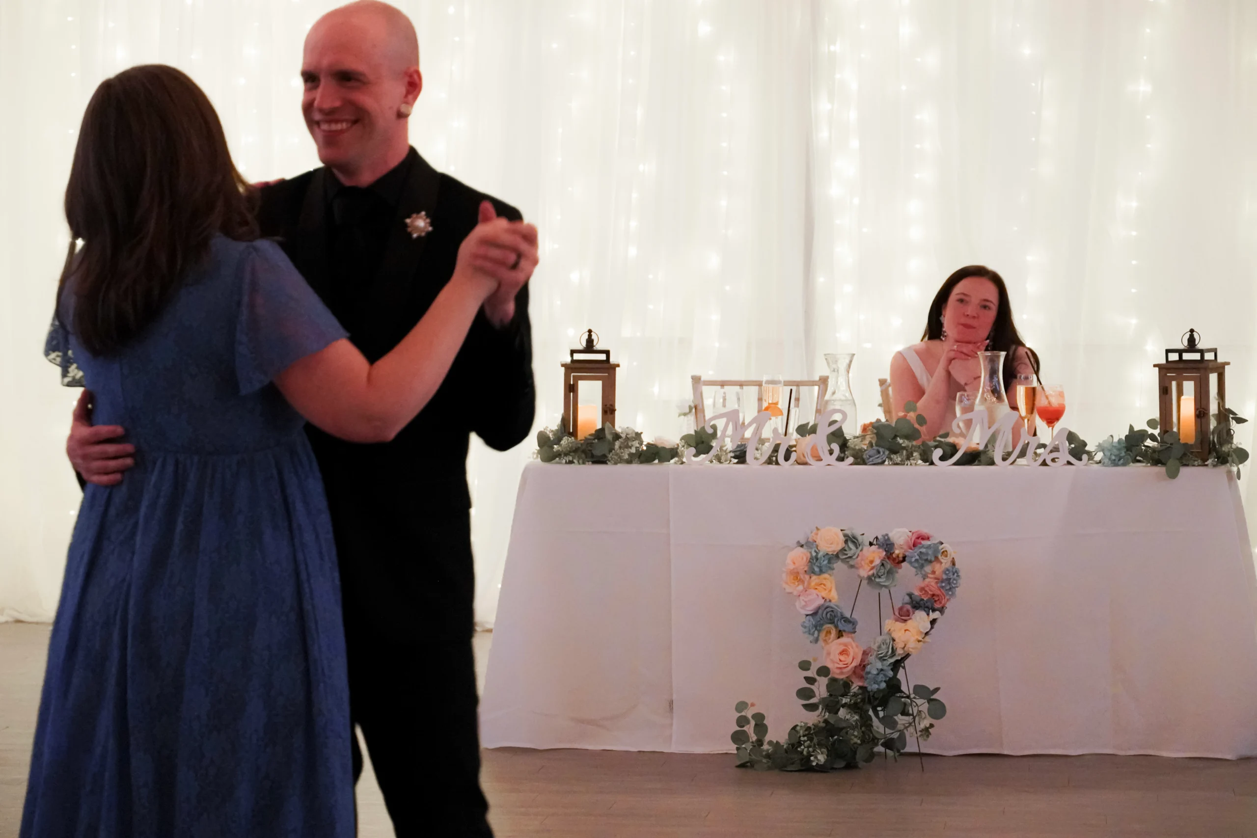 The groom dances with the mother of the bride during the first dances at the wedding reception. The groom is smiling in a black tuxedo. The mother has her back facing the camera, and her hand in the grooms hand. The bride is lovingly gazing at the groom dancing with her mom, from a table in the back of the room with flowers on it, that also says "Mr. & Mrs." in white cursive writing. There is a candle and lantern at each corner of the table, giving a nice candlelit aesthetic. There are elegant white fairy lights shining and glistening behind them.
