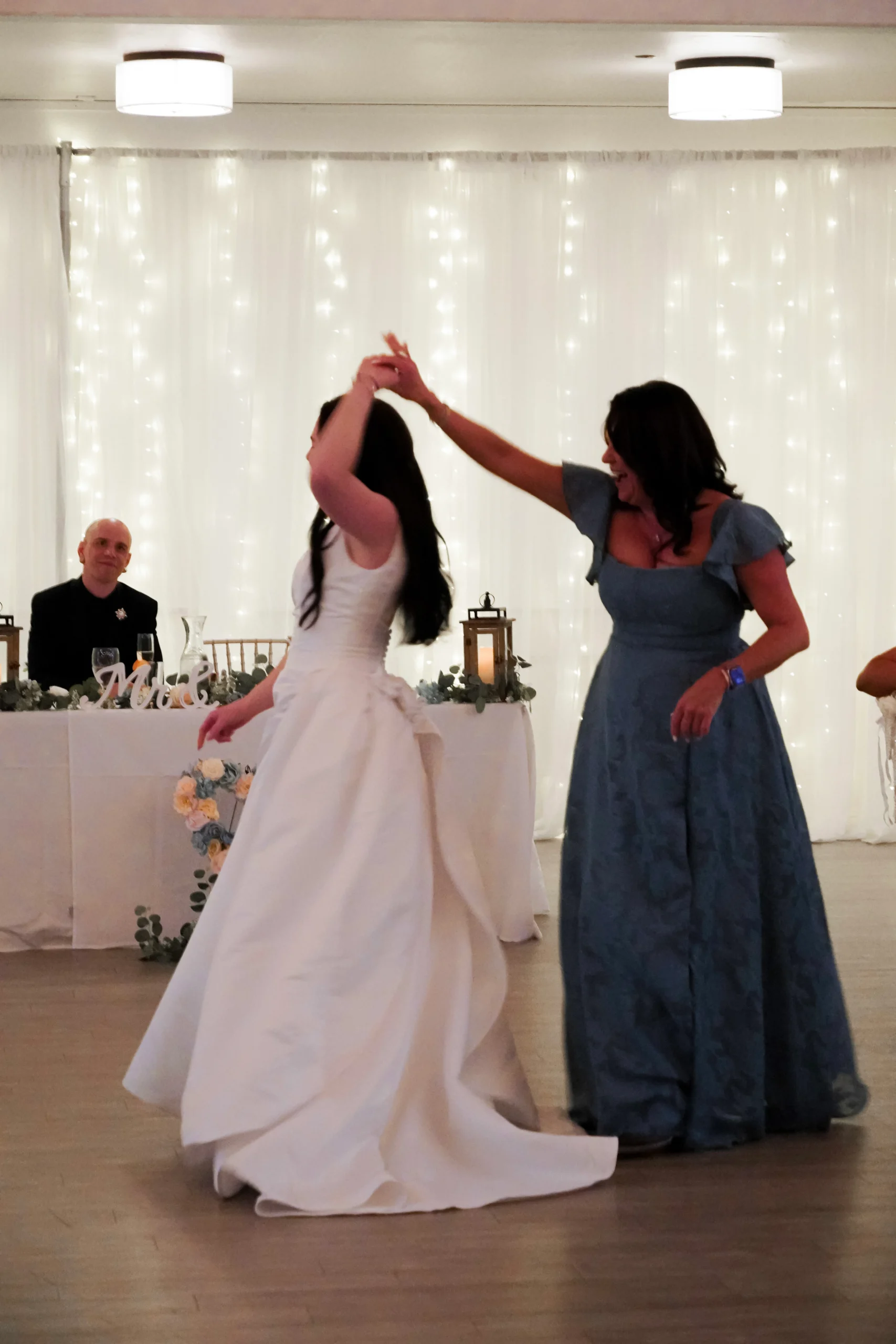 A bride and her mom dance at the wedding reception. The mom twirls the bride, giving a nice silhouette portrait of the bride showing off her wedding reception dress. the background is sparkling and magical. The groom is smiling at his bride dancing with her mom.
