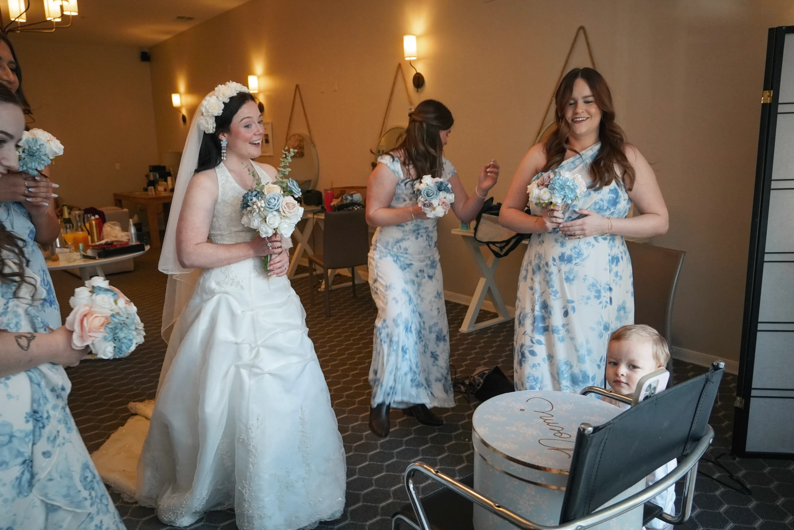 Bride and bridesmaids make a TikTok in the women's dressing room before the ceremony. All of them are smiling, holding bouquets of flowers, and there is a baby watching. The lighting is perfect, airy, but also warm and magical.
