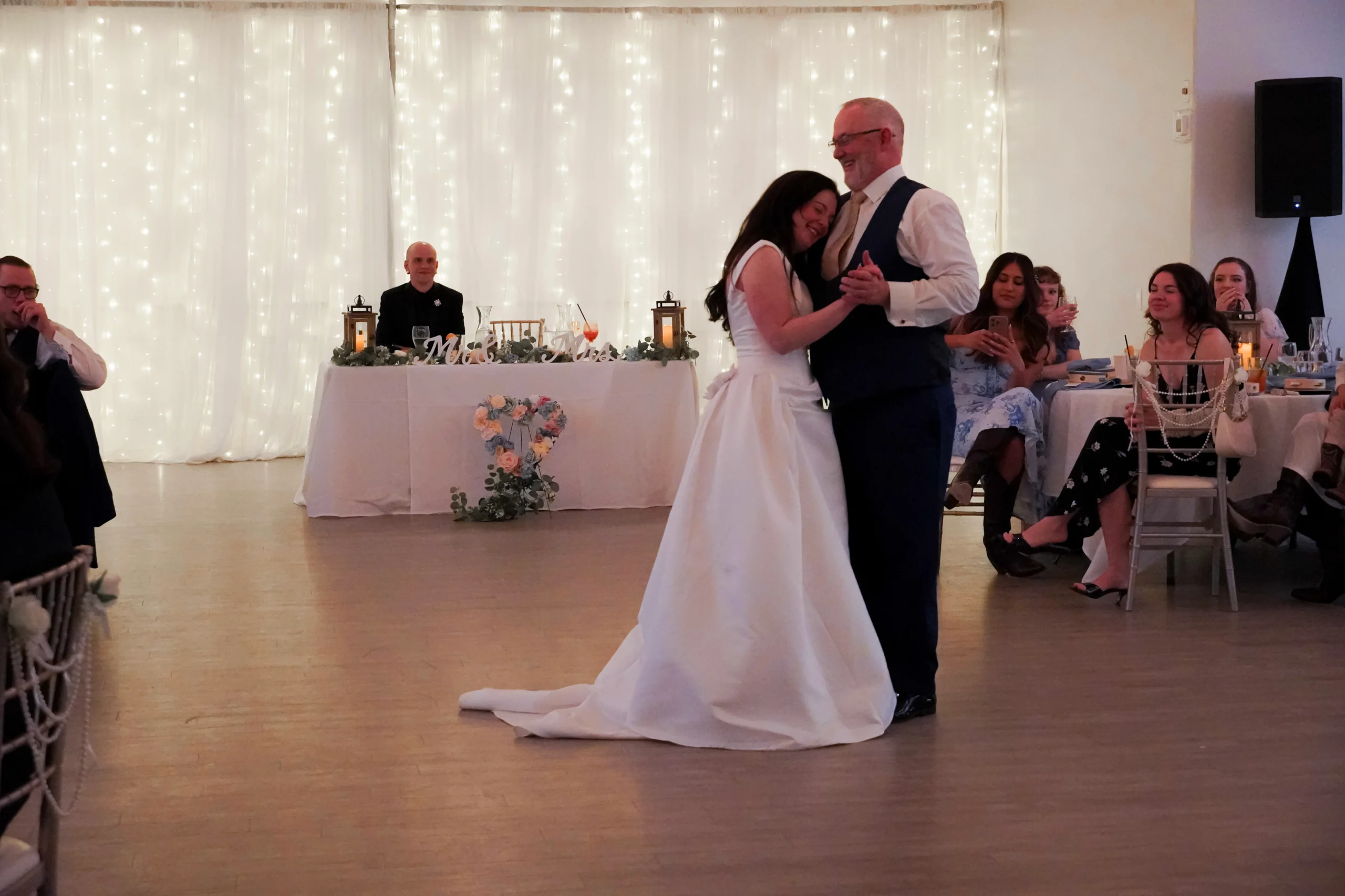 A father dances with his daughter after she gets married, at the reception. Her new husband looks at them and smiles from a table that says "Mr. & Mrs." The guests watch the father and daughter dance from their tables. The bride and her father are smiling. She is leaning her head on his chest. The walls are glittering with lights. It looks like a very happy moment.