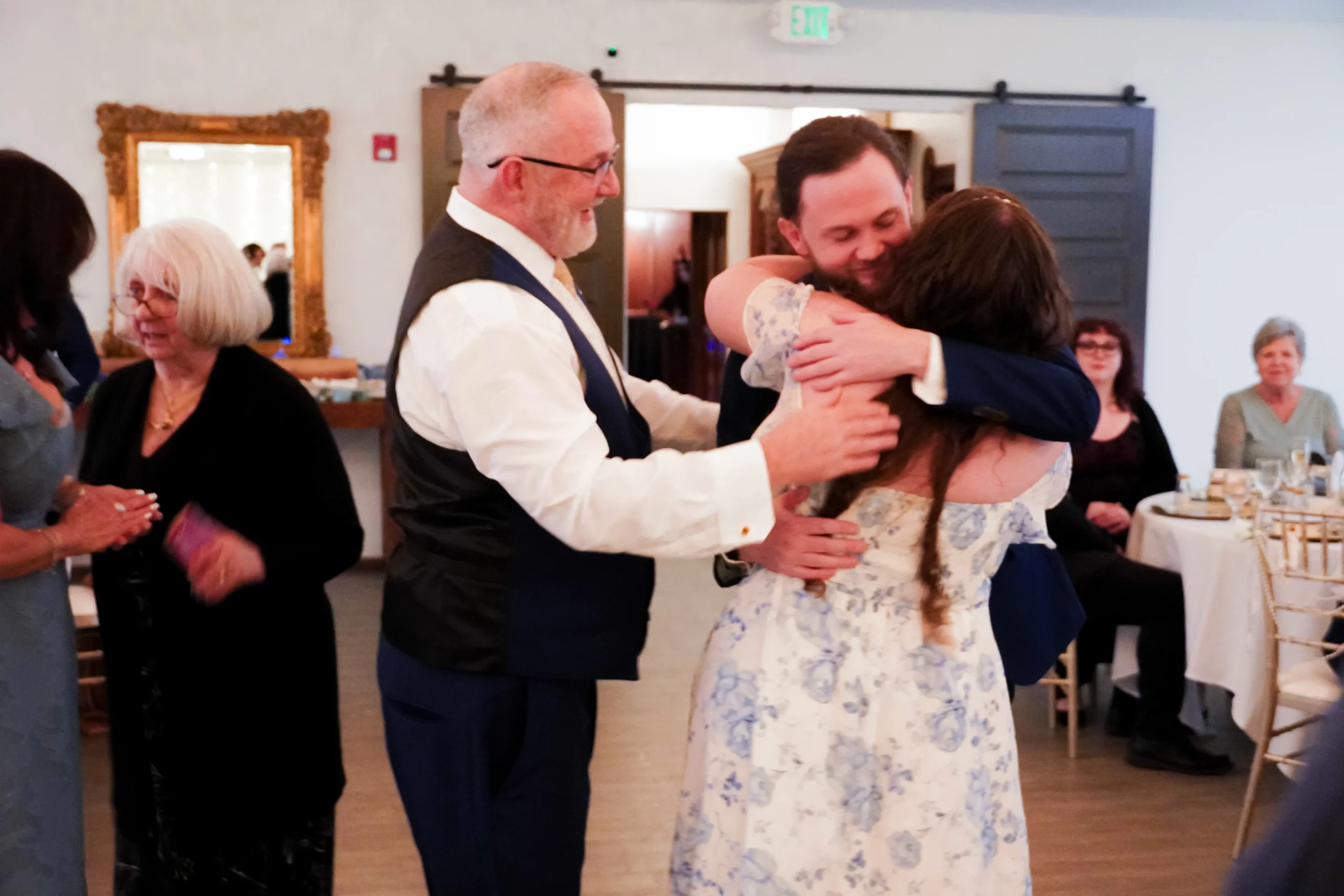 Dad pats his sons soon-to-be-wife on the back while she hugs her new fiancé. The three of them look extremely happy.