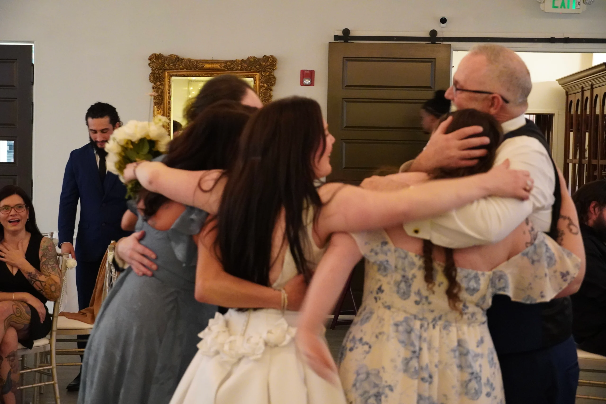 father of the bride, mother of the bride, brother of the bride, and soon-to-be sister-in-law of the bride all enjoy a happy family group hug. Wedding guests in the background are happy for them. They all look very excited.