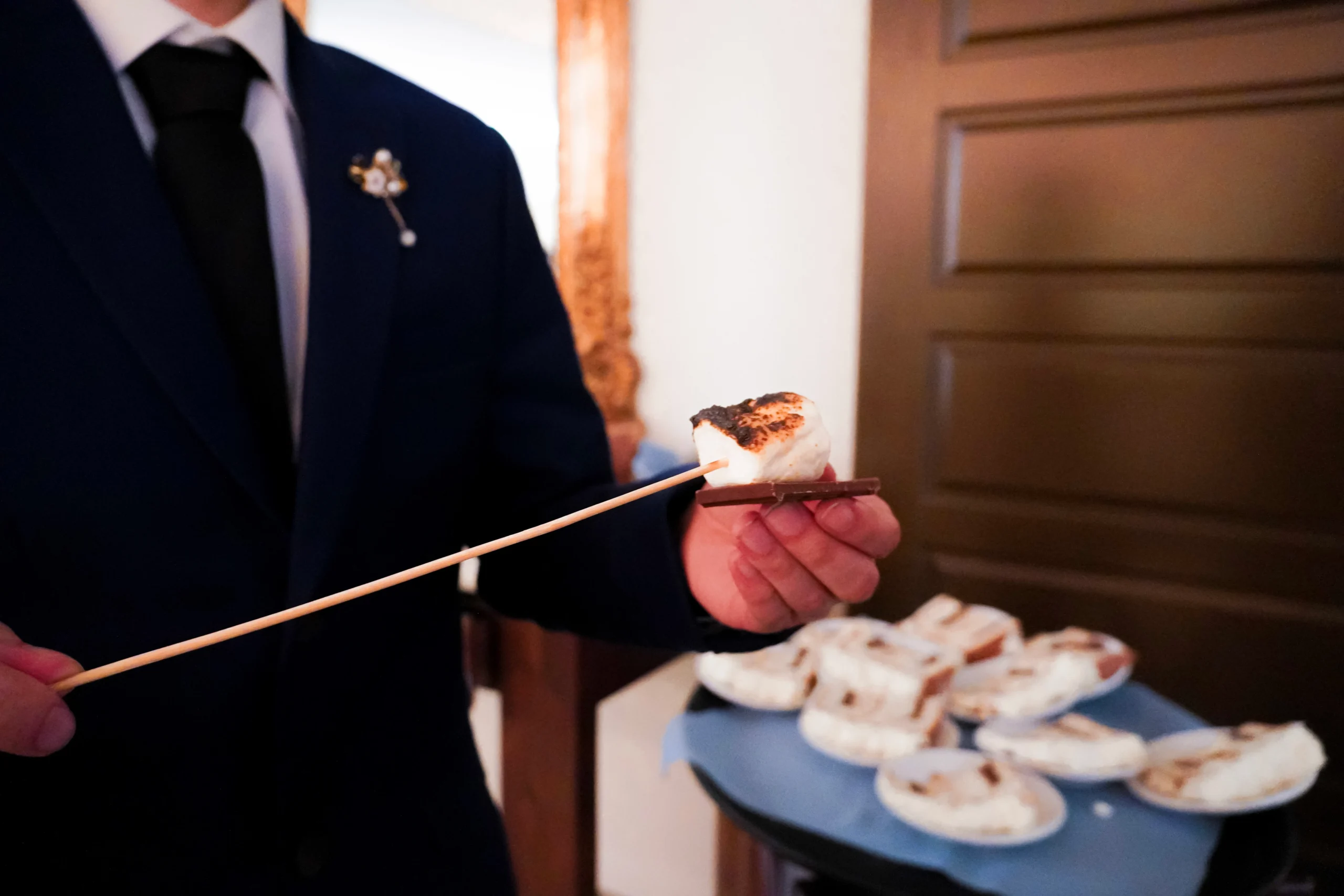 This is a detail shot of the best man showing off a roasted marshmallow on a chocolate bar that he got at the smores bar during wedding reception. He is also standing in front of sliced pieces of wedding cake for the guests. He is wearing a dark blue suit and black tie