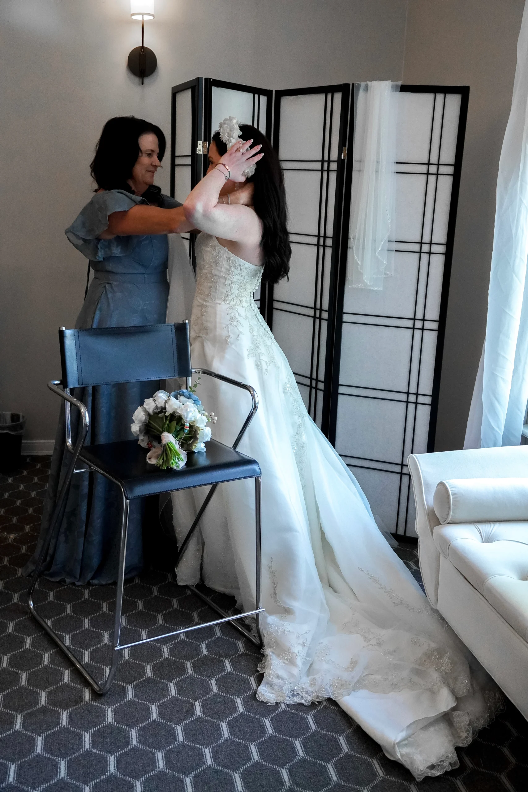 Mother of the bride helps bride get ready in her changing room. The mother is helping tie flowers in her hair that go under the veil. The brides bouquet of flowers is sitting on a chair next to her. The brides dress is white, floral, and sparkly. The bride and her mom are happily looking at each other.