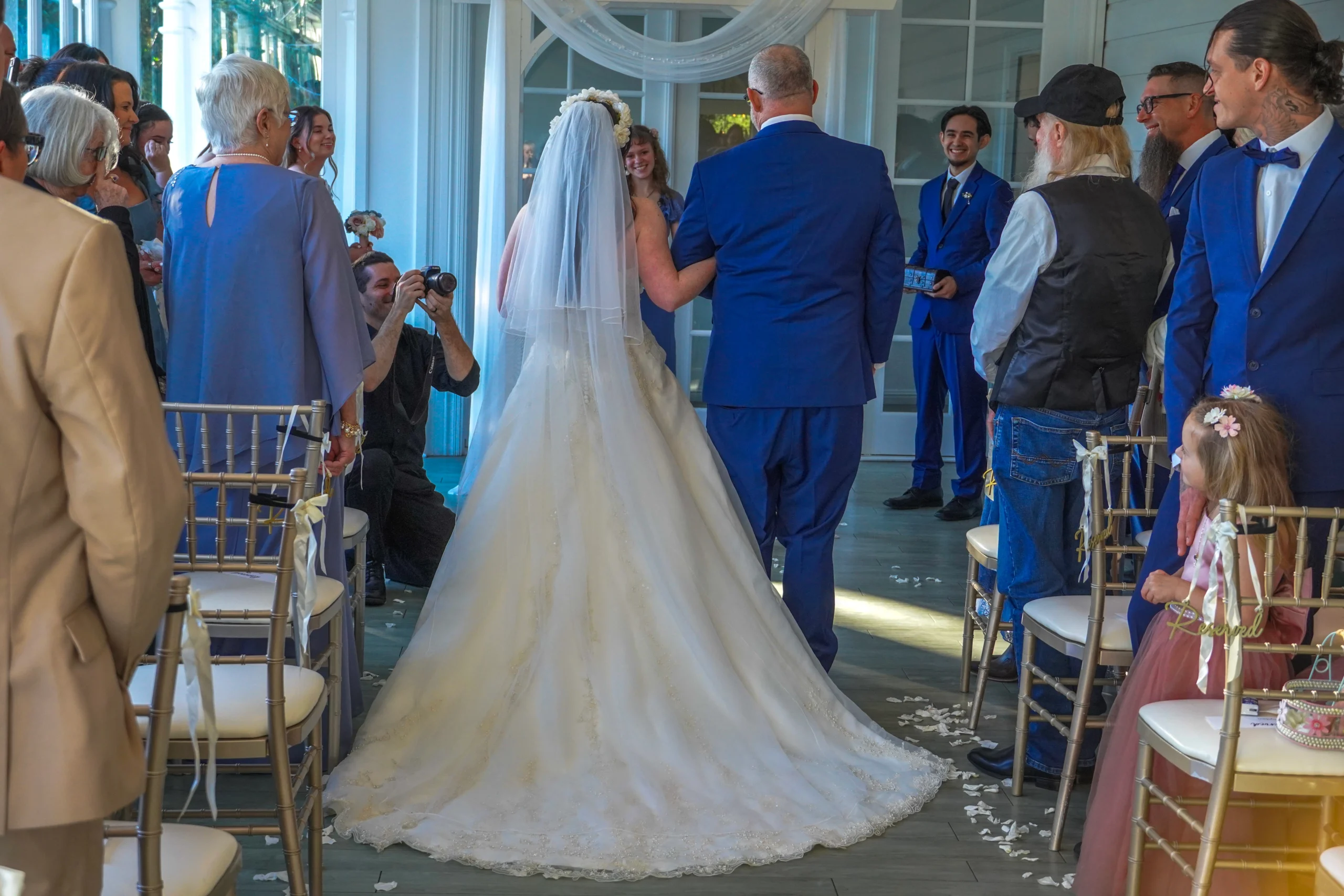The father walks his daughter down the aisle to get married. The bride has a beautiful back of the dress spread out perfectly behind her. A flower girl looks at the bride like she's beautiful. A photographer captures the moment from the front. The best man is facing them and widely smiling. Everyone in the audience is also smiling and happy.