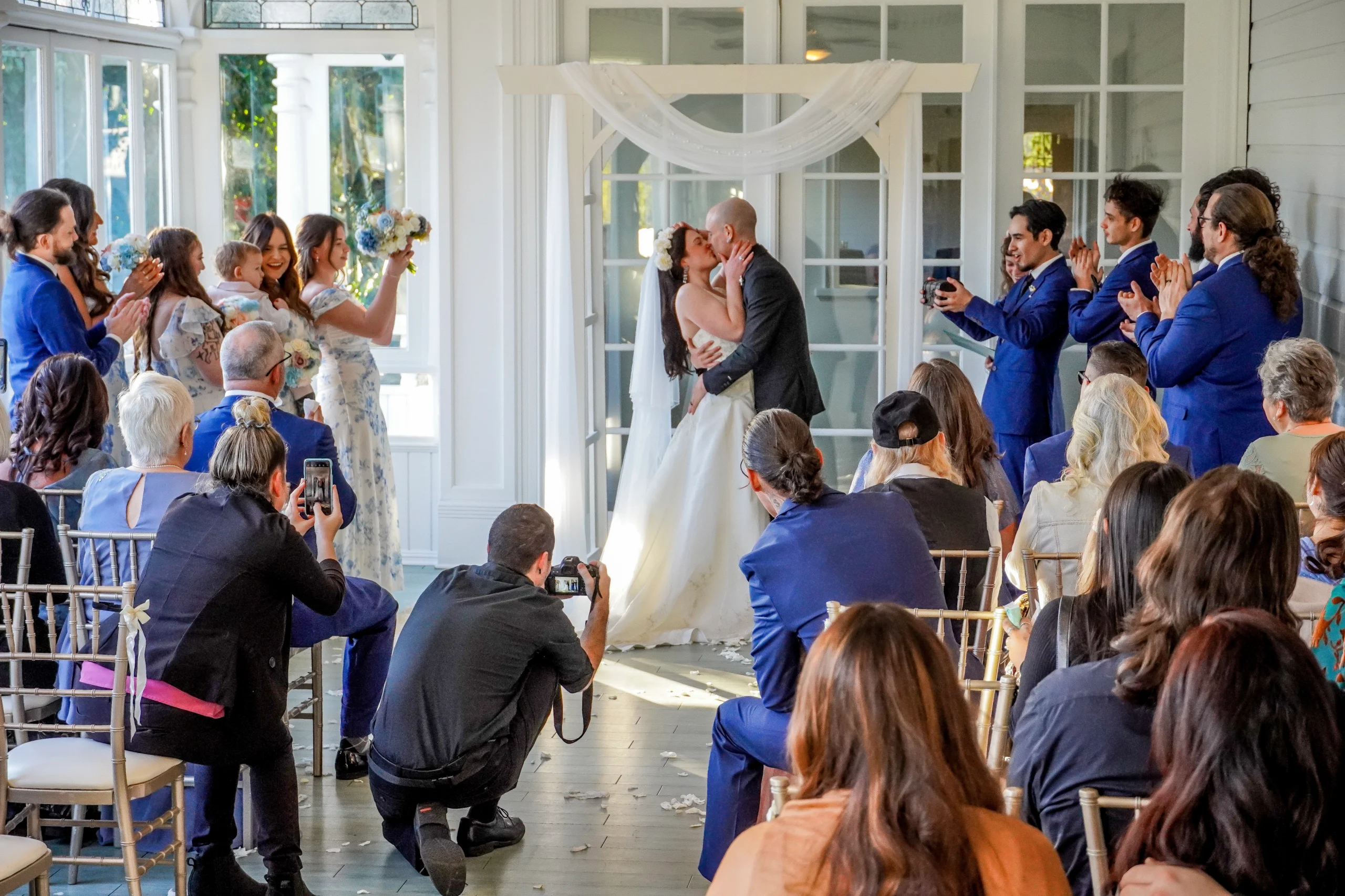 Photographing a beautiful moment of the couples first kiss as husband and wife. The groom kisses the bride during the wedding ceremony. Everyone claps and cheers around them. You can see all their friends and family. A wedding planner and photographer take amazing photos of the couple kissing. The vibe is light and airy spring colors.