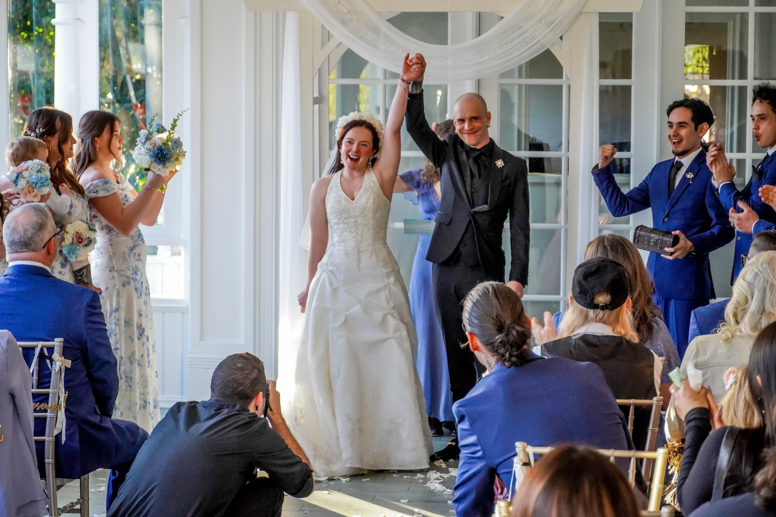 The couple is down the aisle and just married. They are smiling, holding hands, and holding up the hands they are holding in a "we did it!" moment. The people in the crowd are cheering.