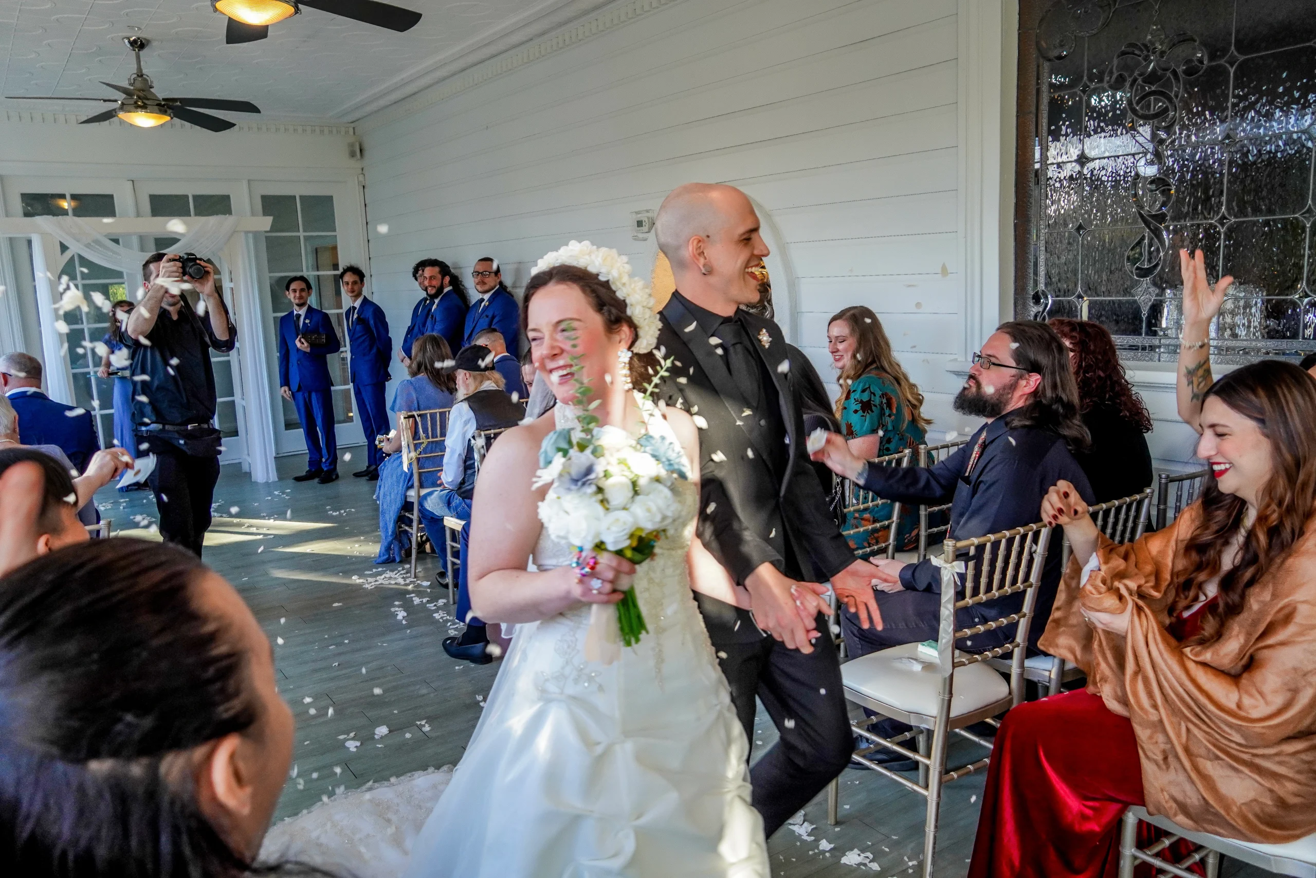 The bride and groom walking down the aisle exiting the wedding ceremony while their friends and family throw flower pedals at the happy couple. The beautiful bride is holding her flower bouquet and is wearing her white dress that sparkles. She is smiling and looking at her guests. The groom is in a classic black suit and tie while also smiling at his guests. There is a photographer taking photos behind them and groomsmen smiling at them leaving. Flower pedals are in the air.