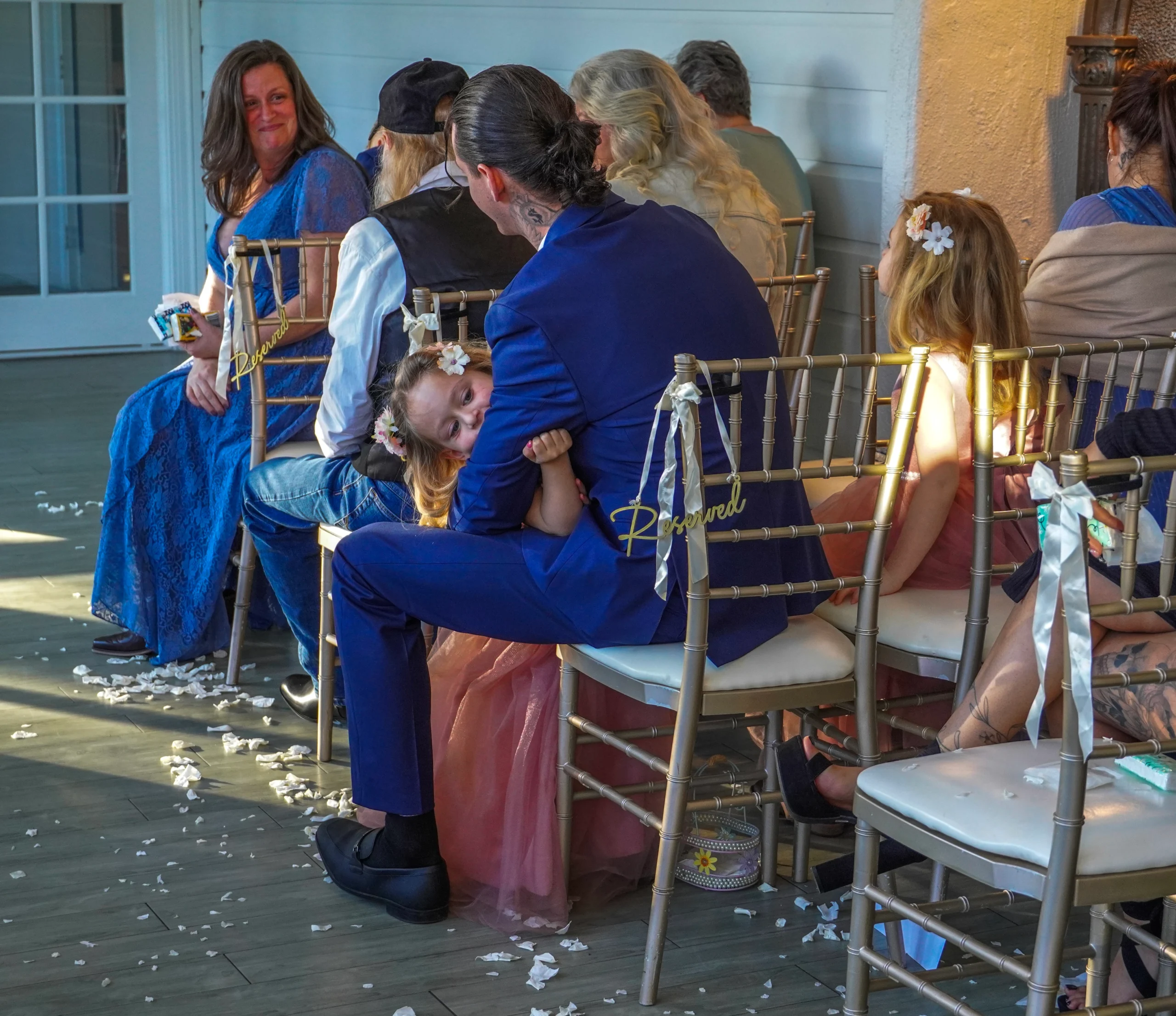 A candid photograph of wedding guests sitting in their seats at the wedding ceremony. There are white flower pedals on the ground and bows on the back of the chairs. Two of the guests are smiling at each other, looking so happy. The flower girls look intrigued but bored, as kids would be. One flower girl is hanging onto her dad and the other flower girl is looking at the aisle.