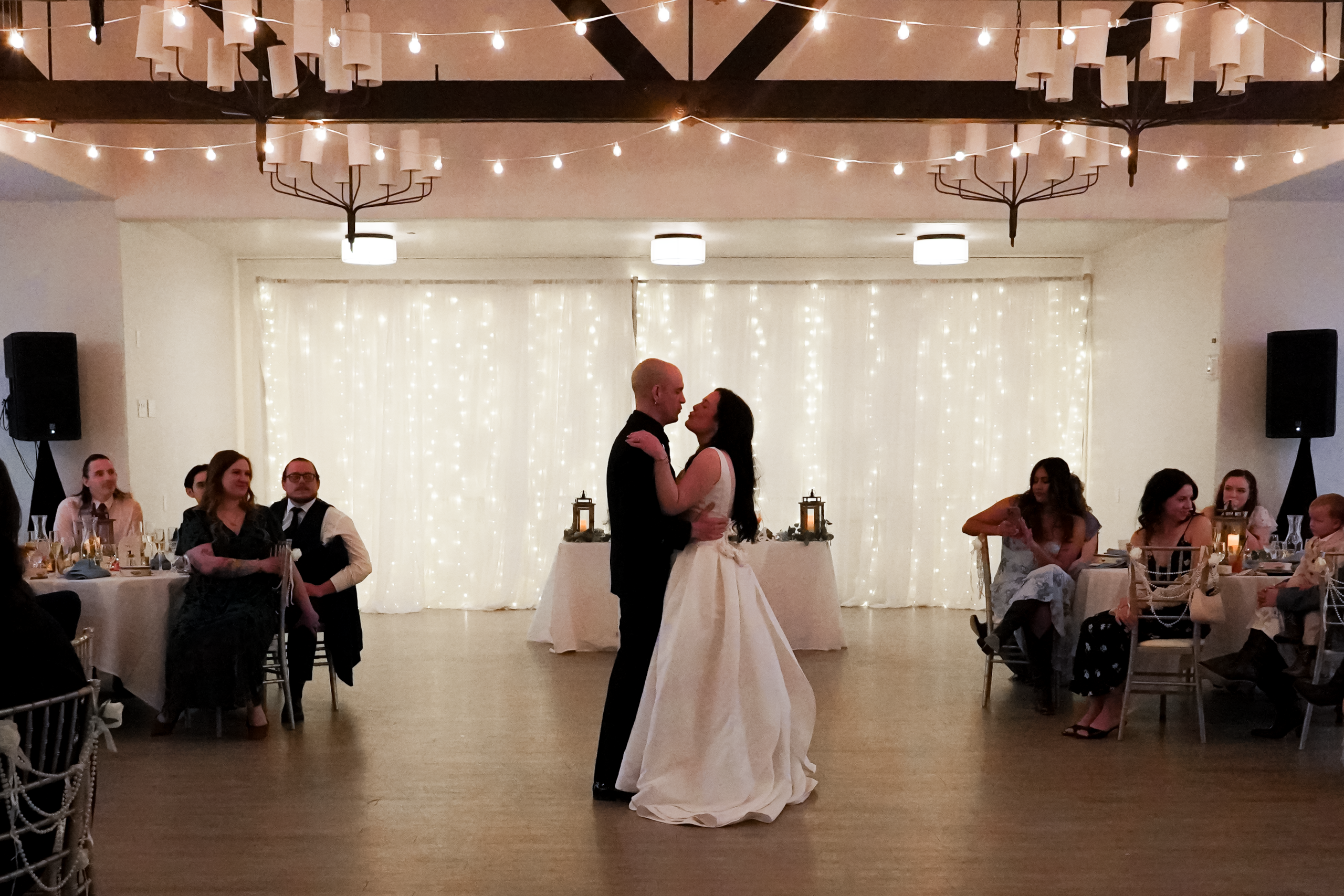 straightened Bride and groom dance their first dance. They are looking at each other, his hands on her waist and her hands on his shoulders, and look like they are about to kiss. They are in the center of the room, with their wedding guests watching them in awe from their dinner tables. The room is dimly lit with twinkling warm white lights above and behind them. It looks like the perfect ballroom wedding photo.
